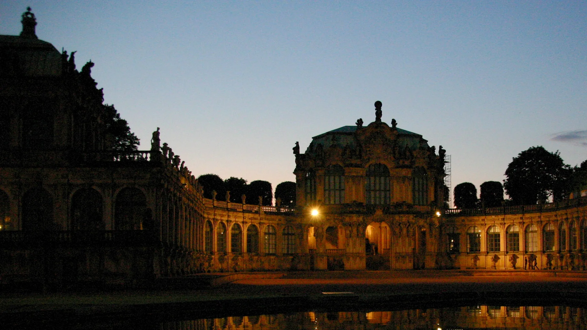 Wallpavillon i Dresden Zwinger i skumringen: en barok pavillon med oplyste buevinduer, buede arkader på begge sider og et stille bassin, der spejler bygningen.
