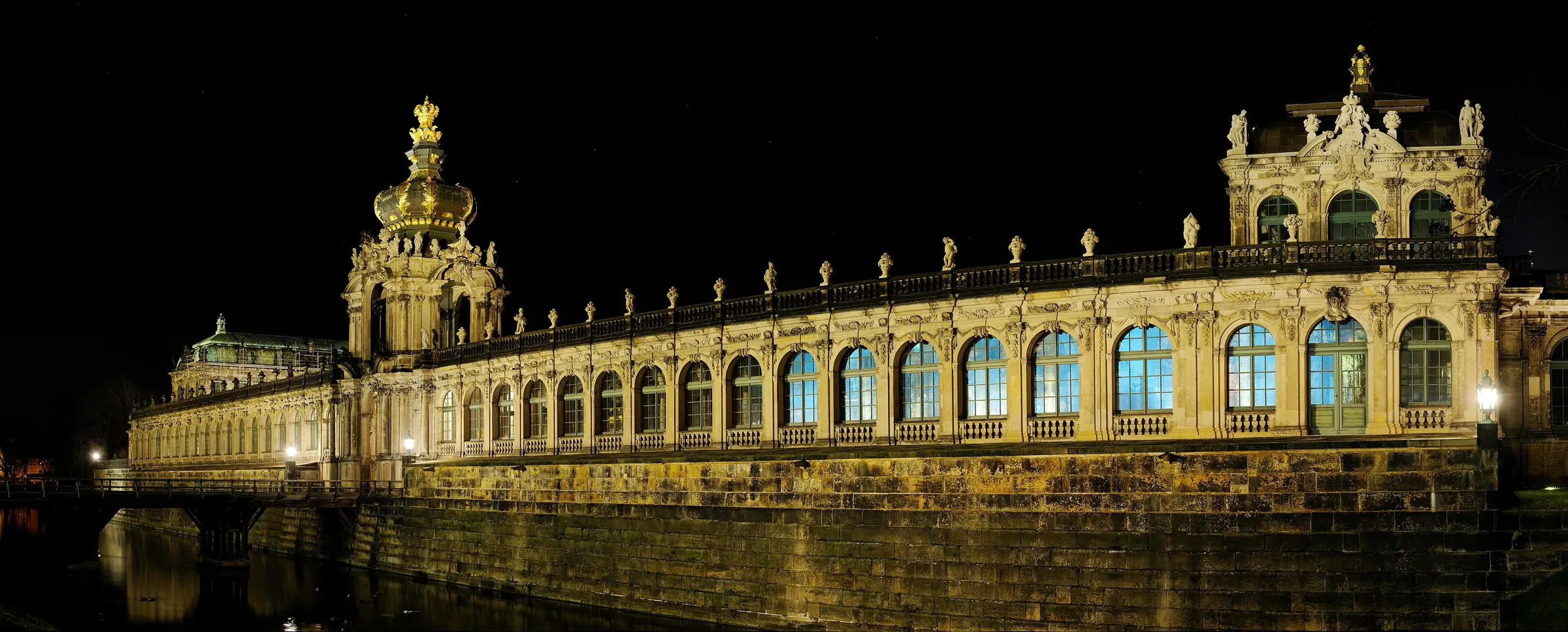 The Zwinger at night: the Crown Gate and the long gallery lit in gold above a dark moat, with a second pavilion at the right-hand end.