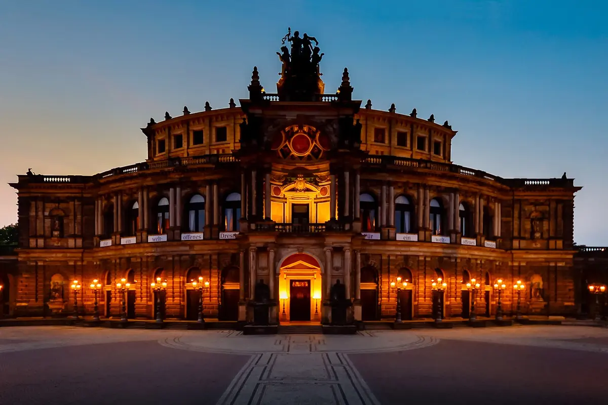 Semperoper i tusmørket med sin oplyste indgangsarkade, rækker af lamper og en skulpturgruppe over taglinjen.